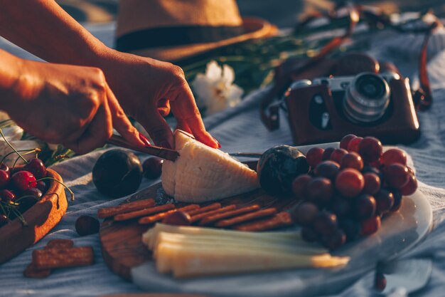 woman slicing cheese on wooden cutting board with cheese and fruits on it and camera, hat and flowers in beach.