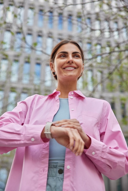 Free photo woman smiles gently checks time on wrist watch waits for someone comes on meeting wears pink shirt poses against city building