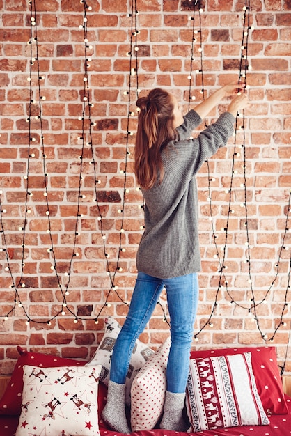 Free photo woman standing on bed and hanging christmas lights