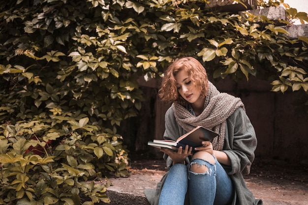 Free photo woman in street wear sitting on board and attentively reading book near plant