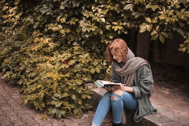 Free photo woman in street wear sitting on board and reading book near plant