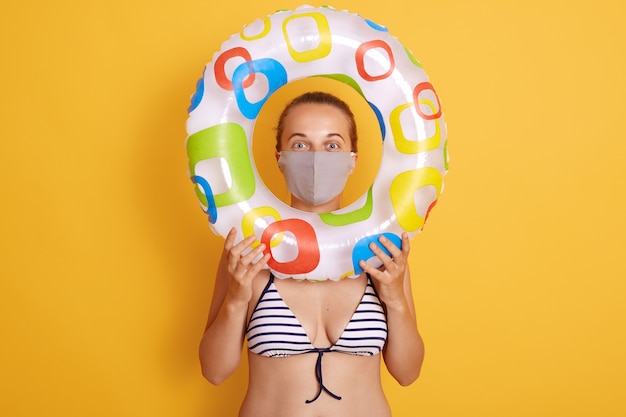 Free photo woman in striped swimsuit looking through rubber ring, wearing hygienic face mask to prevent virus on resort beach, rest and summer vacation with means of protection her health.