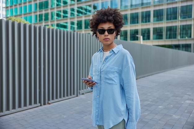 woman strolls with telephone outside wears sunglasses and blue shirt strolls during leisure in city urbanity uses mobile application chats in social media concentrated away