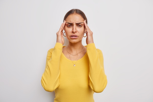 Free photo woman suffers from headache keeps hands on temples frustrated of failure grimaces from painful ache needs painkillers wears casual yellow jumper on white