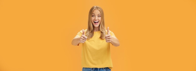 Free Photo woman supports with raised thumbs up and amused cheerful smile showing positive attitude expressing like on concept or idea giving approval posing happy and delighted against orange background