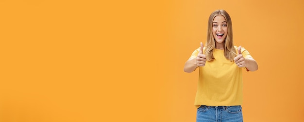 Free photo woman supports with raised thumbs up and amused cheerful smile showing positive attitude expressing like on concept or idea giving approval posing happy and delighted against orange background