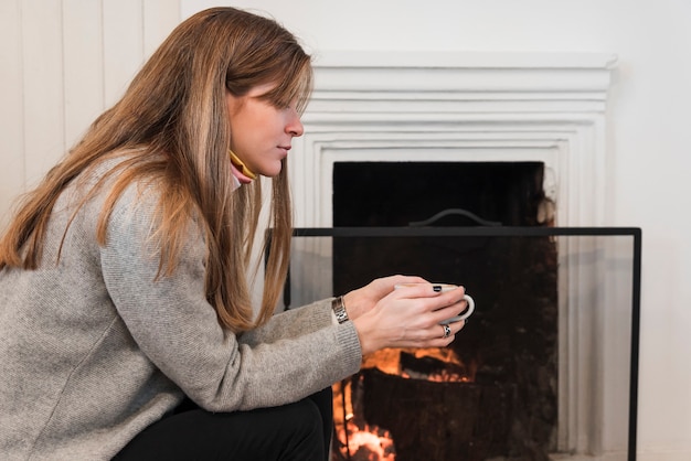 Woman in sweater drinking tea near fireplace