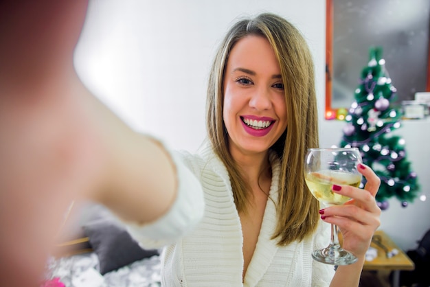 Woman take selfie on phone with Christmas tree hold glass of vine, celebrating New Year. christmas concept - young woman taking selfie photo near decorated christmas tree