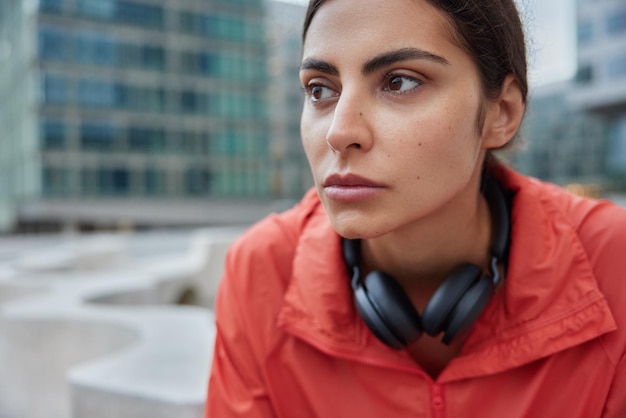 Free photo woman takes break after intensive cardio workout wears windbreaker uses headphones for listening music focused forward poses outdoor