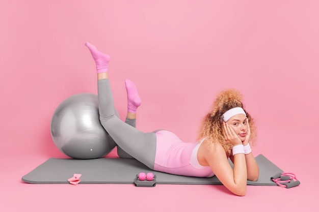 Free photo woman takes rest after fitness training lies on comfortable mat keeps legs on fitball has bored expression dressed in sportsclothes has workout