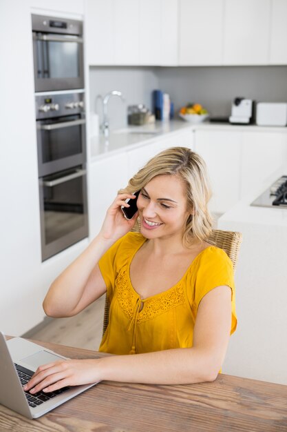 Woman talking on mobile phone while using laptop in kitchen