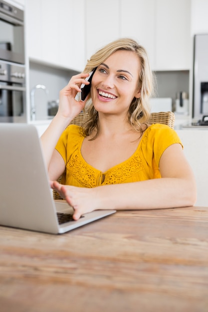 Woman talking on mobile phone with laptop in kitchen