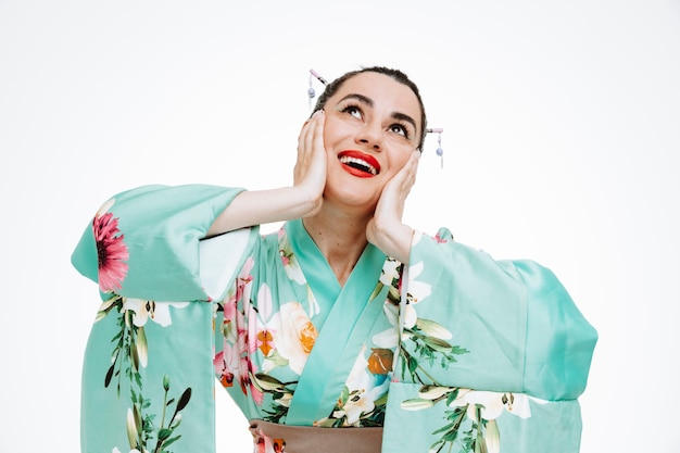 Free Photo woman in traditional japanese kimono looking up happy and surprised dreaming holding hands on her cheeks smiling broadly on white