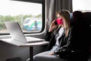 Free photo woman traveling by train and talking on the phone while wearing medical mask and working on laptop