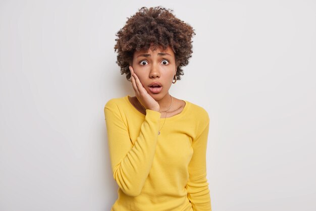 woman trembles from fear stares with speechless expression keeps hand on cheek cannot believe in something amazing wears casual yellow jumper isolated on white