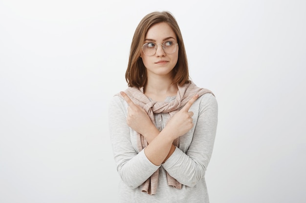 Woman trying chose which product better. Unsure relaxed and smart creative woman with short brown hair in glasses looking right while crossing arms on chest pointing in both sides uncertain