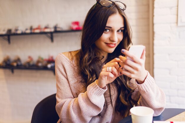 Woman typing write message on smart phone in a modern cafe. Cropped image of young pretty girl sitting at a table with coffee or cappuccino using mobile phone.