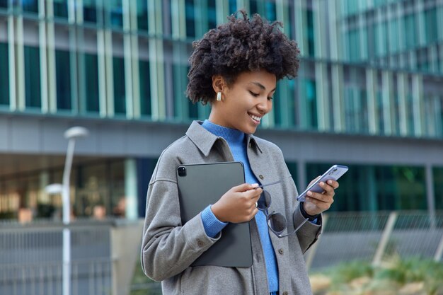 woman uses modern technologies in city holds mobile phone and digital portable tablet smiles gladfully as reads pleasant text message wears stylish clothes