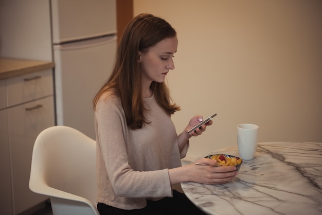 Woman using mobile phone while having breakfast in kitchen