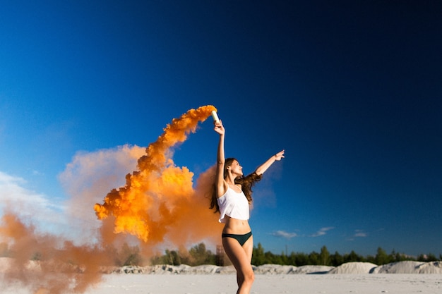 Woman walks with orange smoke on white beach under blue sky