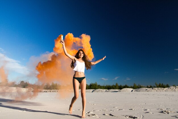 Woman walks with orange smoke on white beach under blue sky