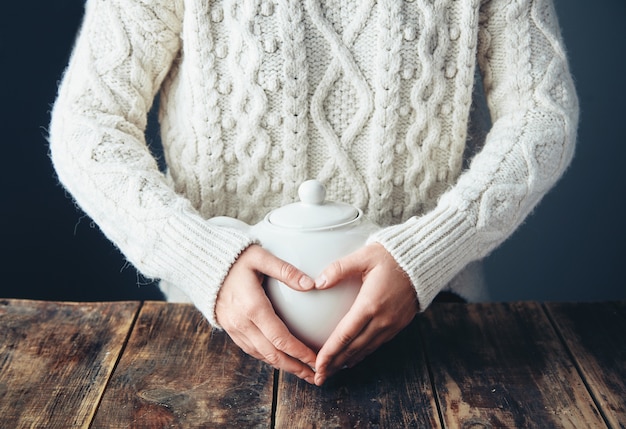 Woman in warm sweater holds hands on big white teapot with tea in heart shape. Front view, grunge wooden table. anfas, no face.