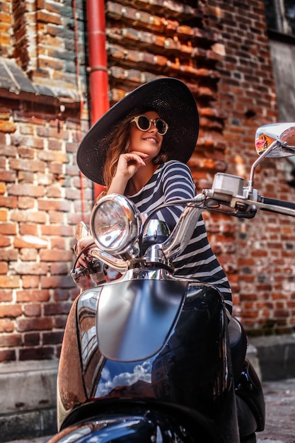 Woman wearing dress and hat sitting on a black classic Italian scooter on an old street in Europe.