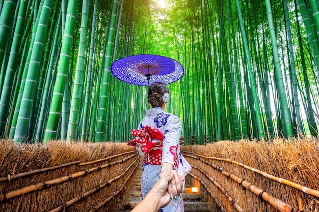 Woman wearing japanese traditional kimono holding man's hand and leading him to Bamboo Forest in Kyoto, Japan.