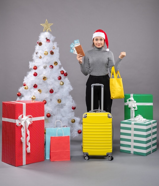 Free photo woman wearing santa claus hat with luggage next to christmas tree