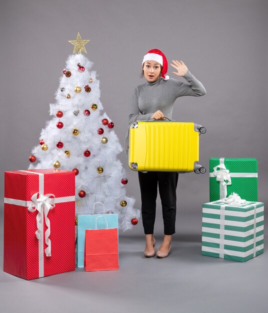 Woman wearing Santa Claus hat with luggage next to Christmas tree