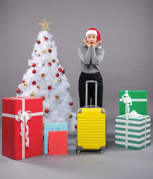 Free photo woman wearing santa claus hat with luggage next to christmas tree