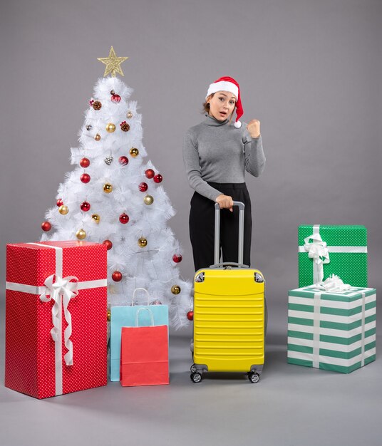 Woman wearing Santa Claus hat with luggage next to Christmas tree