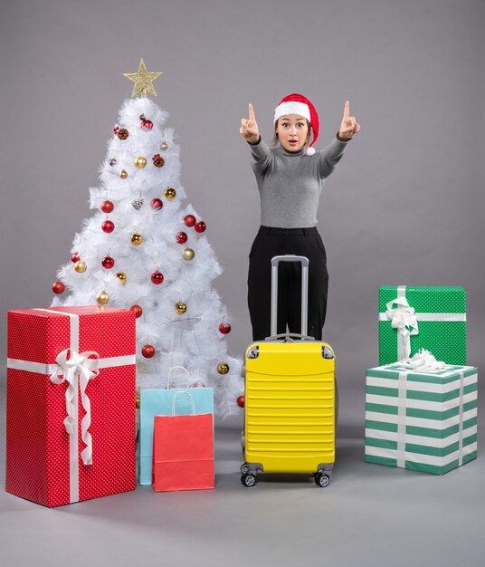 Woman wearing Santa Claus hat with luggage next to Christmas tree