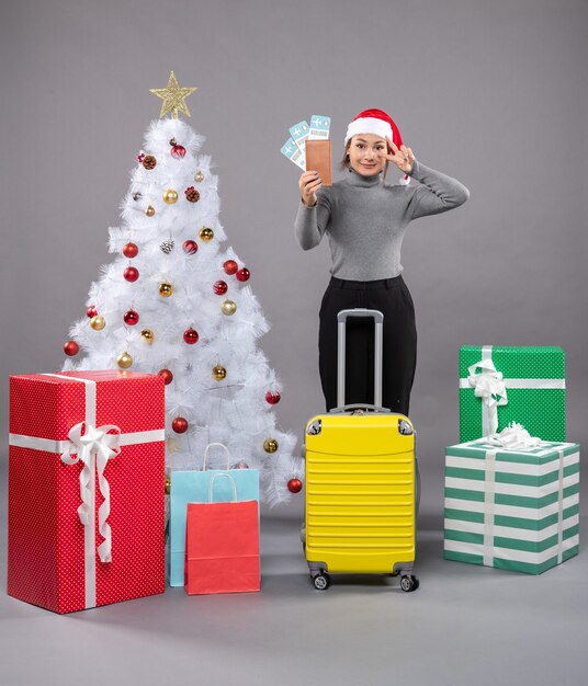 Woman wearing Santa Claus hat with luggage next to Christmas tree