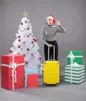 Free photo woman wearing santa claus hat with luggage next to christmas tree