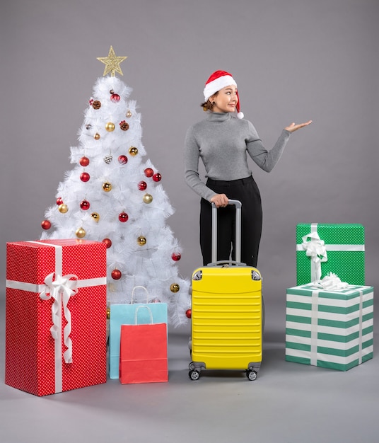 Free photo woman wearing santa claus hat with luggage next to christmas tree