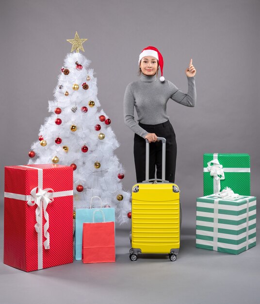 Free photo woman wearing santa claus hat with luggage next to christmas tree