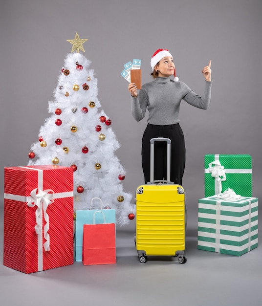 Free photo woman wearing santa claus hat with luggage next to christmas tree