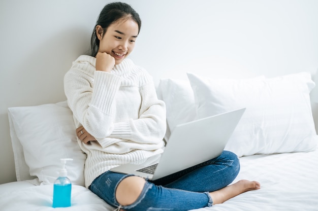 A woman wearing a white shirt on the bed and playing laptop happily.