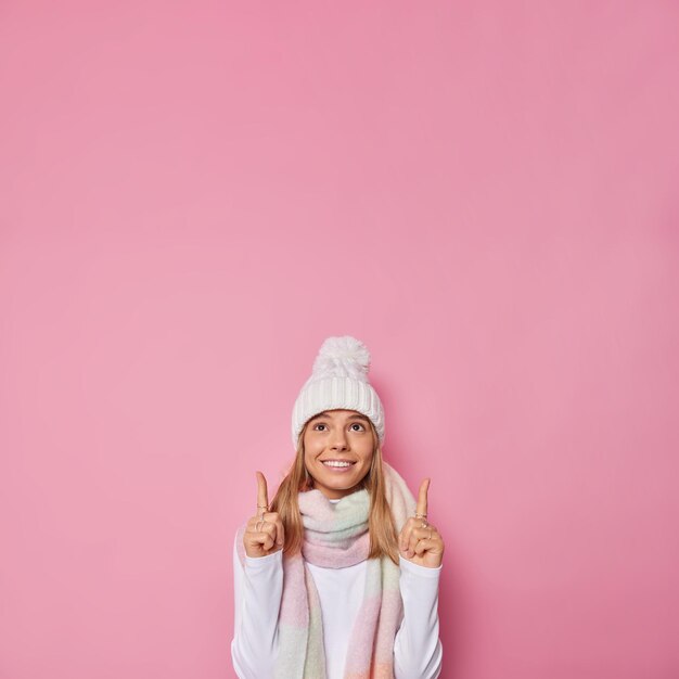 woman wears winter hat and scarf around neck poses against pink points above at blank copy space