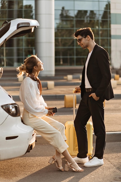 Woman in white blouse beige pants sits in car trunk and talks to her boyfriend Brunette man and blonde lady speak and pose with yellow suitcase near airport