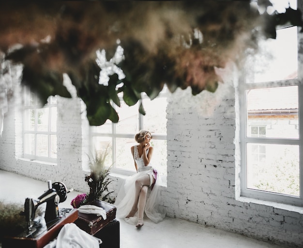 Free photo woman in white clothes sits on the windowsill in a room with flowers and sewing machine