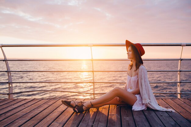 woman in white dress sitting by the sea on sunrise in romantic mood wearing red hat