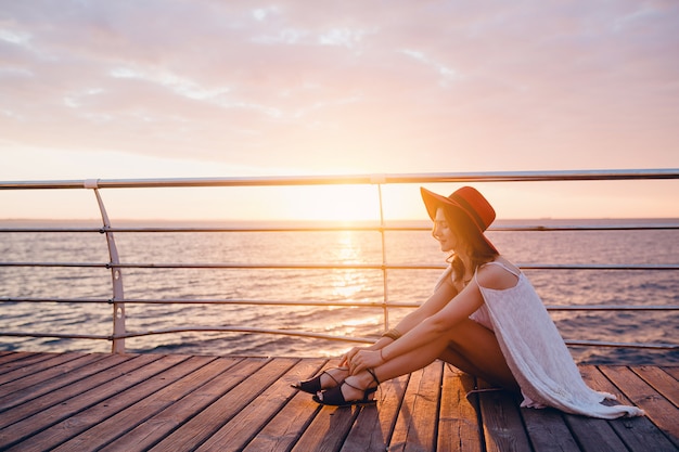 woman in white dress sitting by the sea on sunrise in romantic mood wearing red hat