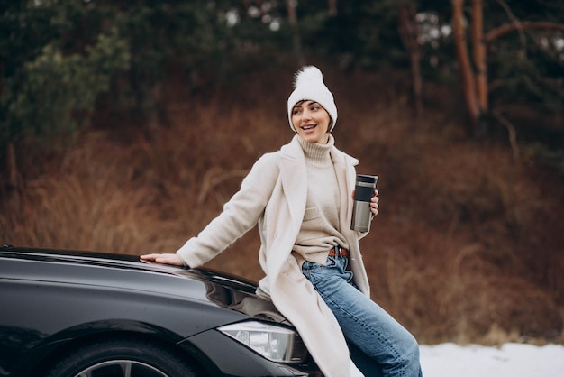 Woman in winter time sitting on car hood and drinking coffee