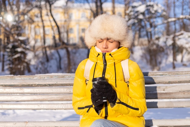 Free Photo a woman in winter in warm clothes in a snow-covered park on a sunny day sits on a bench and is freezing from the cold, is unhappy in winter, holds coffee alone