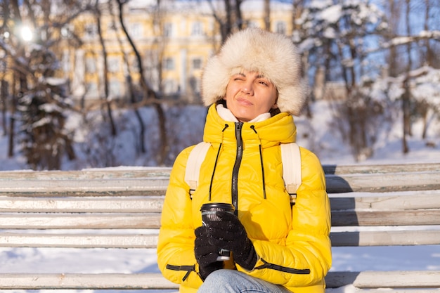 Free Photo a woman in winter in warm clothes in a snow-covered park on a sunny day sits on a bench and is freezing from the cold, is unhappy in winter, holds coffee alone