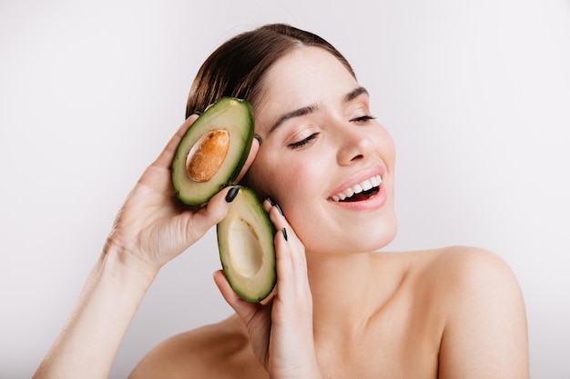 Woman with bare shoulders cute smiles posing with avocado useful for skin on white background