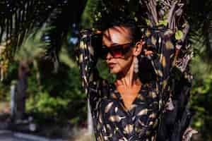 Free photo woman with bronze make up wearing black golden dress and sunglasses in tropical landscape