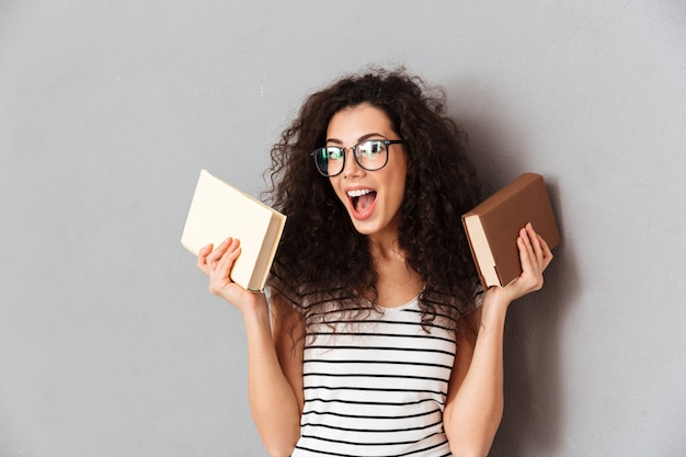 Free Photo woman with brown curly hair being student in university posing with interesting books in hands taking pleasure in education isolated over grey wall